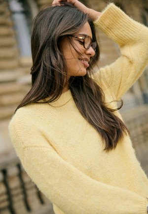 Jeune femme aux longs cheveux foncés portant des lunettes écaille et un pull jaune, souriant avec une main dans les cheveux en extérieur.