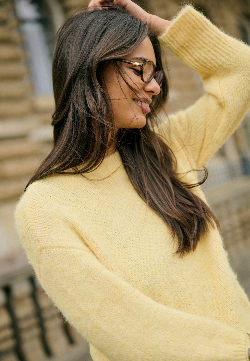 Jeune femme aux longs cheveux foncés portant des lunettes écaille et un pull jaune, souriant avec une main dans les cheveux en extérieur.