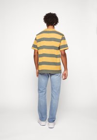 Tričko s krátkým rukávem, pruhované v hořčicové žluté a olivové zelené, s kulatým výstřihem a volnějším střihem. Kombinováno se světle modrými džíny.