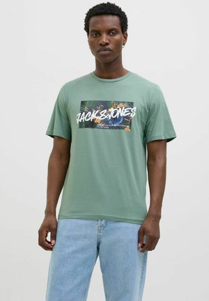 Grøn bomulds t-shirt med et centralt graphic design med tropiske mønstre og fed tekst. Klassisk rund hals og korte ærmer. Let tekstur.