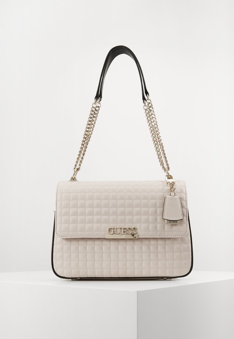 Sac bandoulière matelassé beige avec des bretelles en chaîne argentée, une poignée noire et un logo "GUESS" argenté sur le rabat avant.