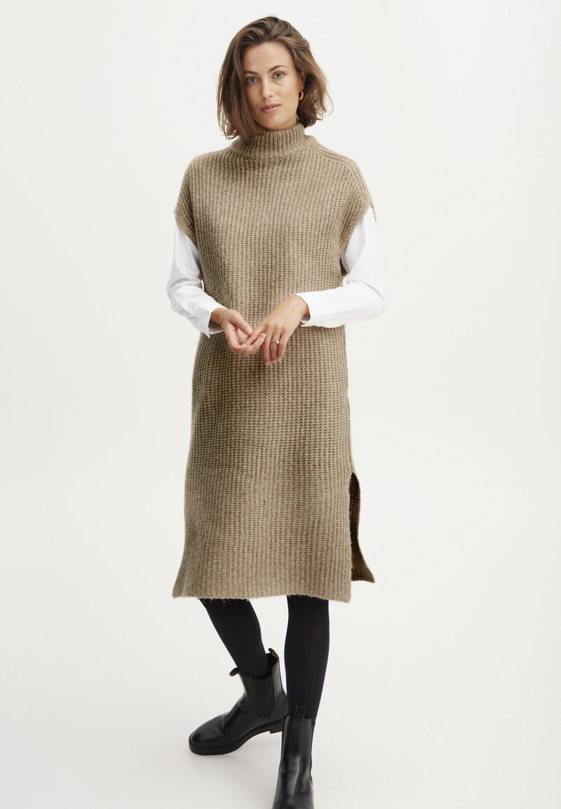 Beige Strickkleid mit hohem Kragen und kurzen Ärmeln, mit Seitenschlitzen, getragen über einem weißen Langarmshirt, kombiniert mit schwarzen Knöchelstiefeln.
