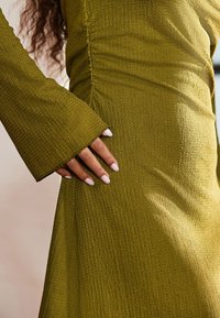 Olivegrünes, strukturiertes Kleid mit Glockenärmeln. Nahaufnahme zeigt zerknitterten Stoff und eine Hand, die an die Seite gelehnt ist, mit zart rosa Nagellack.