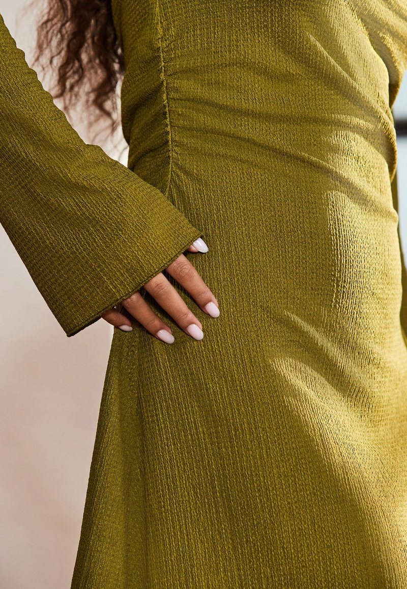 Olivegrünes, strukturiertes Kleid mit Glockenärmeln. Nahaufnahme zeigt zerknitterten Stoff und eine Hand, die an die Seite gelehnt ist, mit zart rosa Nagellack.