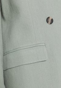 Tecido de blaser verde claro com um acabamento texturizado, apresentando um botão castanho único e um bolso com aba do lado esquerdo.