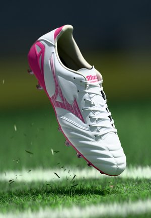 White and pink Mizuno soccer cleat mid-air above green grass with scattered grass blades and white field markings.
