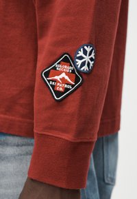 Κόκκινο πουκάμισο με μακριά μανίκια που διαθέτει δύο κεντημένες ετικέτες: μία μαύρο διαμάντι με τη φράση «COLORADO ROCKIES SKI PATROL CO» και ένα μπλε νιφάδα χιονιού.