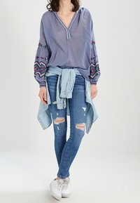 Blusa bordada de manga larga en azul claro con escote en V; combinada con jeans skinny desgastados en azul y zapatillas blancas, camisa de mezclilla atada a la cintura.