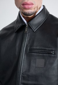 Veste en cuir noir avec une fermeture éclair, col pointu, poche zipée à l'avant et un écusson logo carré sur la poitrine. Texture lisse.