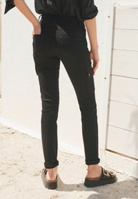 Mustat slim-fit farkut, joissa on sileä pinta, sivutaskut ja käärityt lahkeet, yhdistettynä tummiin sandaaleihin hiekkaisella pinnalla.