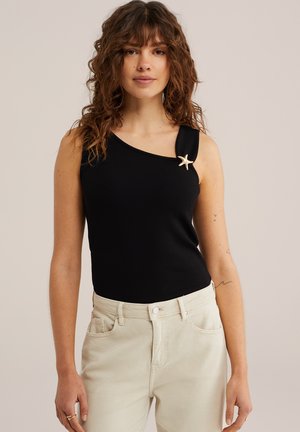 Žena s kučeravými vlasmi nosiaca čierny top na jedno rameno s brošňou v tvare morskej hviezdy a svetlo béžové nohavice, stojaca pred jednofarebným pozadím.