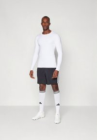 Fehér hosszú ujjú ing fekete rövidnadrággal, fehér focizokni fekete csíkokkal és fehér focicipő; minimalista kialakítás.