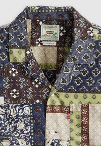 Košulja s kratkim rukavima s patchwork uzorkom koji uključuje mornarsku plavu, smeđu, zelenu i bež cvjetne i geometrijske uzorke; dolazi s prednjim džepom i gumbom.