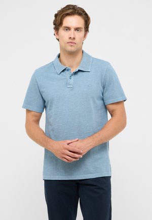 Mann trägt ein hellblaues Polohemd mit kurzen Ärmeln und dunkle Hose, steht mit verschränkten Händen vor einem einfarbigen Hintergrund.