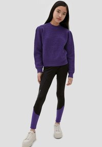 Pull en texture violet, leggings noirs avec des accents et un logo violet, et baskets blanches à plateforme. Accent sur les contrasts de couleurs et de matériaux.
