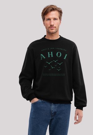 Schwarzer Sweatshirt mit langen Ärmeln und rundem Hals, mit türkisfarbenem Text "AHOI" und Vogelgrafiken. Getragen mit blauen Jeans.