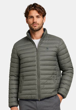 Man wearing a gray quilted zip-up jacket over a white shirt, standing with one hand in pocket against a plain background.