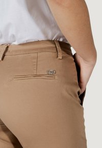 Beige broek met een gladde textuur, slim fit, met een achterzak met een decoratief zilveren embleem en subtiele stiksels.