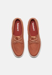 Brązowe płócienne loafersy z białymi gumowymi podeszwami, posiadające zaokrąglony czubek, sznurowadła i czerwone wkładki z nadrukowanym logo Timberland.