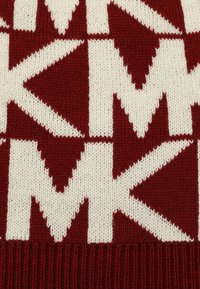 Maglione a maglia con sfondo rosso e grandi motivi bianchi a forma di 'M' interconnessi e frecce, con un texture a coste all'orlo.