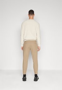 Maglione a maglia beige con scollo rotondo, abbinato a pantaloni tapered color sabbia e scarpe nere, visto da dietro su uno sfondo neutro.