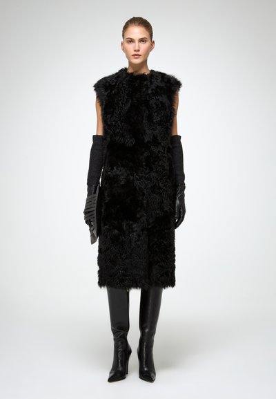 Cappotto in pelliccia sintetica nera con un design senza maniche, abbinato a lunghi guanti neri e stivali neri con tacco a spillo fino al ginocchio, evidenziando texture ed eleganza.
