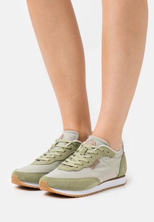 Osoba nosząca jasnoszare i beżowe siatkowe sneakersy z białymi podeszwa środkowymi i gumowymi podeszwami zewnętrznymi na białym tle.
