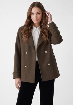 Young woman with long wavy hair wears a dark green double-breasted coat over a white button-up shirt and black pants, posing against a plain background.