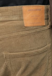 Pantaloni din catifea cord cu un ton khaki, cu nervuri verticale. Prezintă o etichetă din piele maro cu text embosat pe talie. Design simplu al buzunarului din spate.