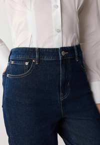 Tamsiai mėlynos džinsinės, aukštu liemeniu, kelnių su auksinėmis siūlėmis, priekiniams kišenėms ir metaliniu mygtuko užsegimu, derinamos su balta marškinių apykakle.