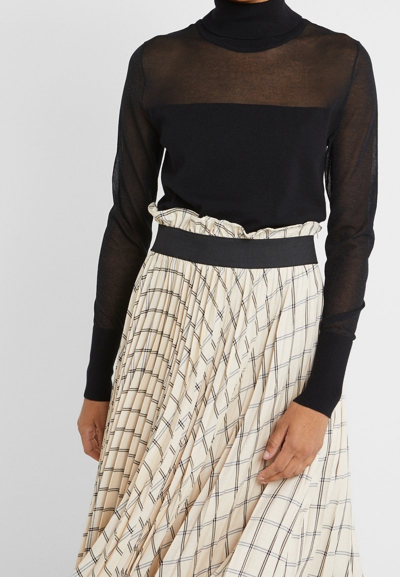 Femme portant un haut col roulé noir transparent à manches longues associé à une jupe plissée beige taille haute avec un motif à carreaux noirs.