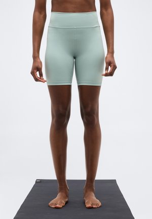 Άτομο που στέκεται ξυπόλητο σε μαύρο στρώμα γιόγκα, φορώντας αθλητικό σορτς σε ανοιχτό πράσινο χρώμα με ψηλή μέση.