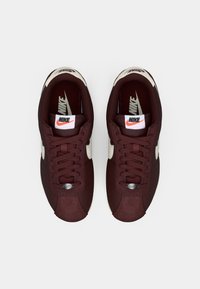 Sneaker Nike color bordeaux con tomaia in tessuto morbido, baffo bianco e lacci piatti. Presenta un occhiello argentato e un colletto imbottito per il massimo comfort.