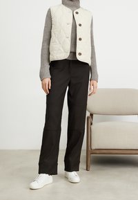 Krémová prošívaná vesta s hnědými knoflíky, nošená přes šedý žebrovaný rolák, doplněná černými širokými kalhotami a bílými teniskami.