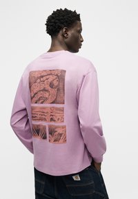 Άνδρας με σγουρά μαλλιά που φοράει πουκάμισο μακρυμάνικο λεβάντας με αφηρημένες καφέ γραφικές πινακίδες στην πλάτη, συνδυασμένο με σκούρο τζιν παντελόνι.