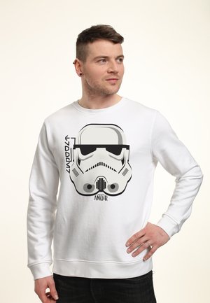 Mężczyzna w białej bluzie z grafiką czarno-białego hełmu szturmowca i napisem Star Wars Andor, pozujący z ręką na biodrze.