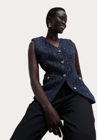 Marineblauw geteerde vest met een V-hals, versierd met goudkleurige knopen en een textuurrijke afwerking. Gecombineerd met zwarte broek.