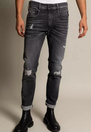 Man wearing rolled-up faded black distressed jeans and black leather boots, standing against a plain background.