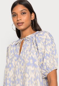 Blusa azul-claro com um padrão texturizado em creme, apresentando um decote em forma de olho de fechadura e mangas curtas bufantes. Com laços no pescoço.
