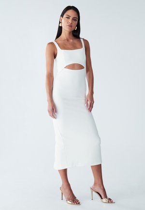 Robe midi blanche avec un décolleté carré, détail découpé à la taille et tissu lisse et texturé. Portée avec des talons hauts dorés.