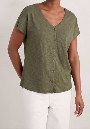 Olivgrünes kurzärmeliges Oberteil mit V-Ausschnitt, mit Knopfleiste vorne, lockerem Schnitt und weichem, strukturiertem Stoff. Kombiniert mit weißen Hosen.