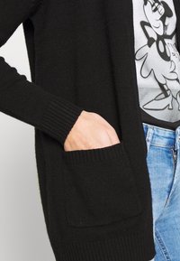 Svart strikket cardigan med ribbede mansjetter og en lomme. Myk tekstur, åpen frontdesign, synlige sømdetaljer, brukt over en grafisk T-skjorte.