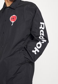 Svart jacka med snap-fram, som har en röd cirkulär logotyp på bröstet och vit "Reebok" text längs ärmen. Slen yta.