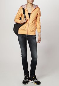 Giacca zip-up arancione chiaro con fodera grigia a contrasto, jeans scuri slim-fit e scarpe da ginnastica nere con accenti colorati, mentre porta una borsa scura.