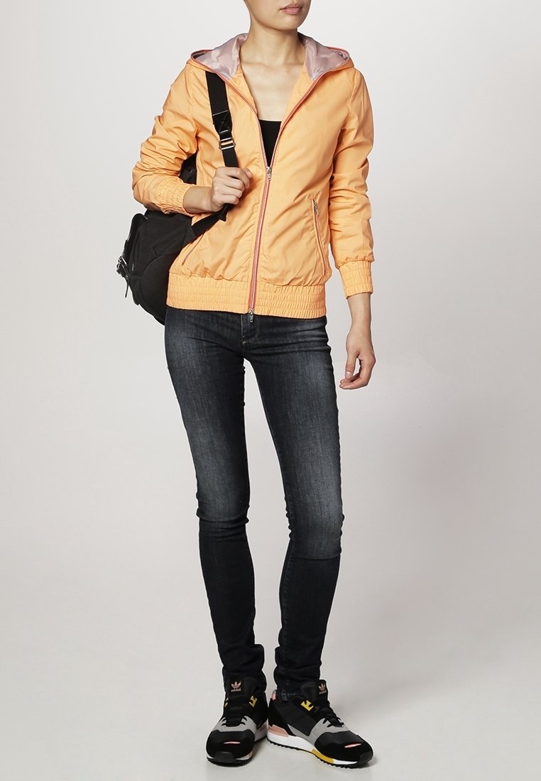 Giacca zip-up arancione chiaro con fodera grigia a contrasto, jeans scuri slim-fit e scarpe da ginnastica nere con accenti colorati, mentre porta una borsa scura.