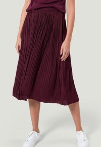 Gonna midi plissè bordeaux con elastico in vita e una texture fluida, abbinata a sneakers bianche su uno sfondo chiaro.