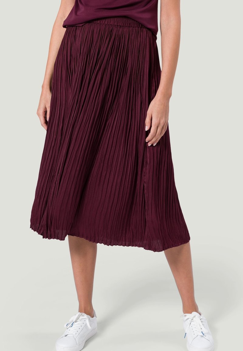 Gonna midi plissè bordeaux con elastico in vita e una texture fluida, abbinata a sneakers bianche su uno sfondo chiaro.