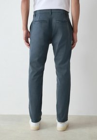 Sötétkék, slim-fit nadrágot viselő személy fehér sportcipővel, egy sima világos háttér előtt állva, hátulról nézve.