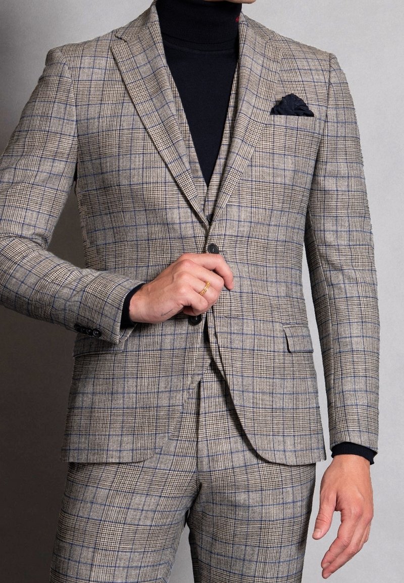 Completo a scacchi beige con righe blu. Vestibilità su misura, rever a punta, chiusura con un solo bottone, abbinato a un dolcevita nero sotto.