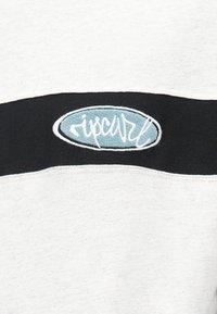 Sivi i crni dukserica s istaknutim plavim ovalnim logotipom s bijelom vezom. Mješavina pamuka, glatka tekstura, dizajn opuštenog kroja.